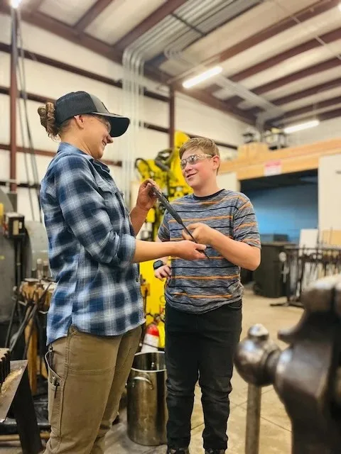 Two people discussing metalwork beside an anvil at Kilroy’s Workshop.